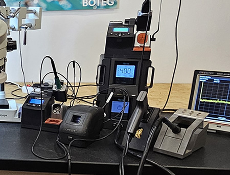 Pohled do dílny, je vidět pracovní stůl s pájkou, odsavačkou, mikroskopem a dalšími nástroji. V pozadí špulíčky s náhradními součástkami.